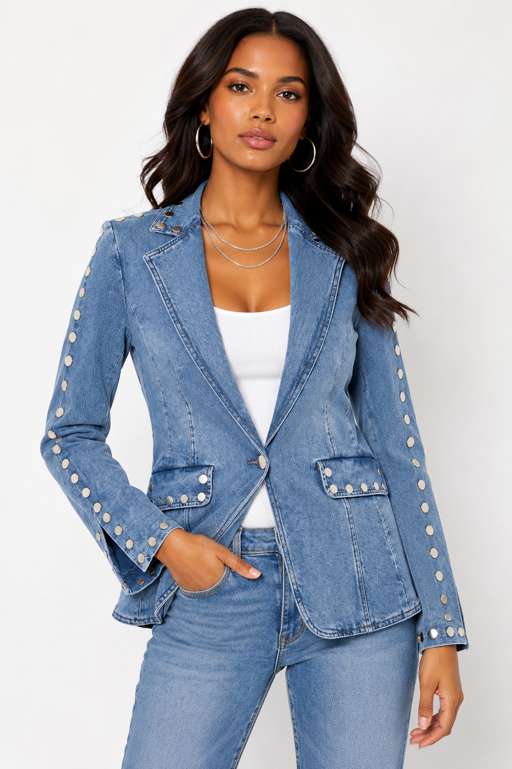 A woman modeling a light blue denim blazer with silver button details on the sleeves, paired with matching blue jeans and a white top, against a plain light gray background.