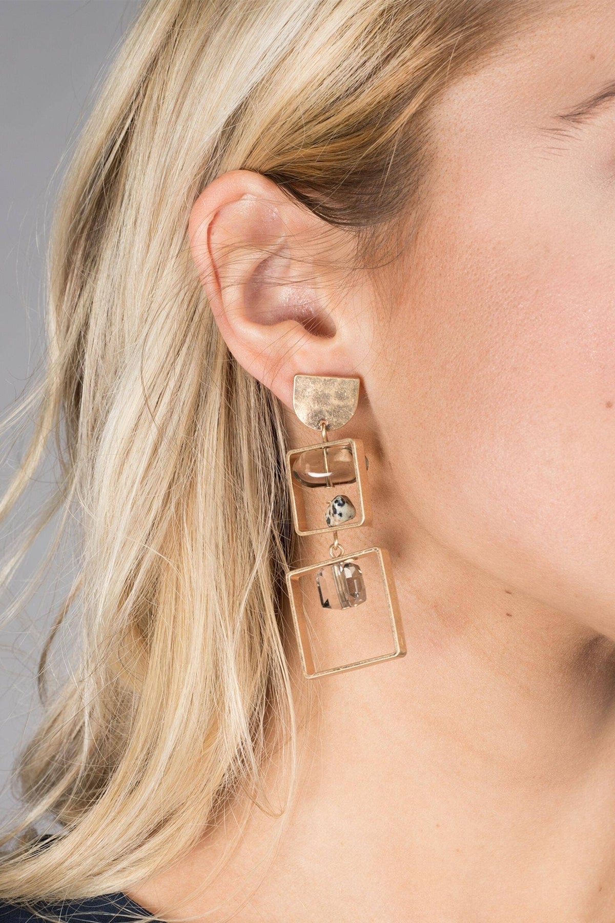 Close-up of a person wearing gold square earrings with clear stones.