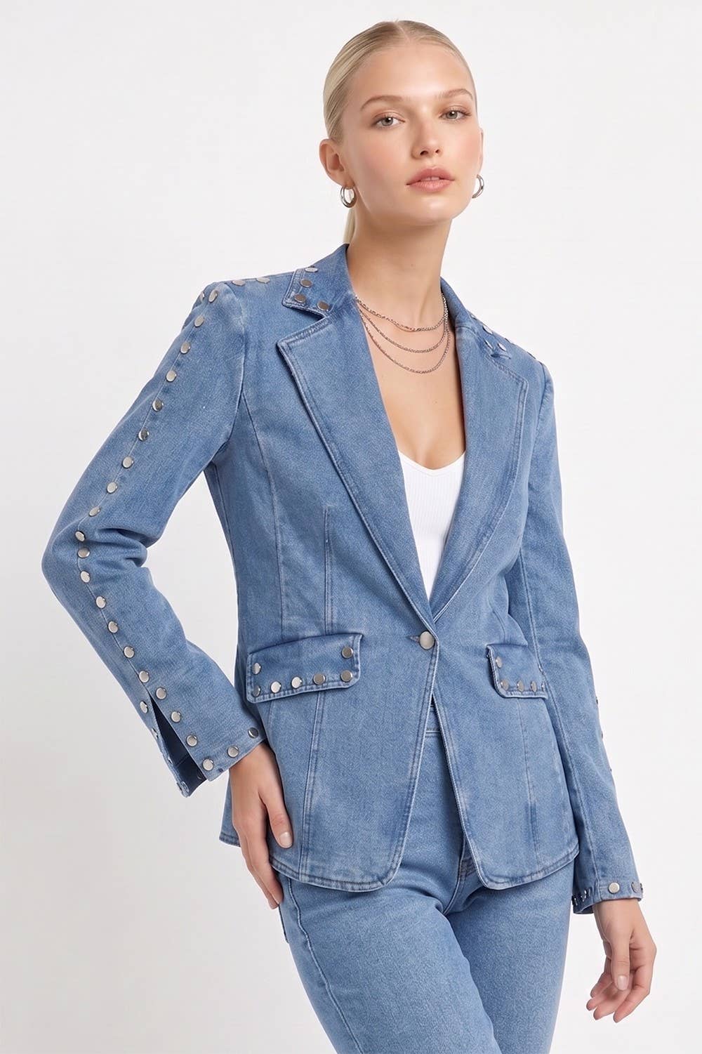 A woman modeling a light blue denim blazer with silver button accents on the sleeves and front pockets, paired with matching blue jeans and a white top, against a plain white studio background.