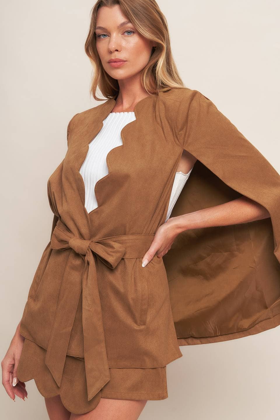 Woman wearing a brown cape over a white top and shorts on a beige background