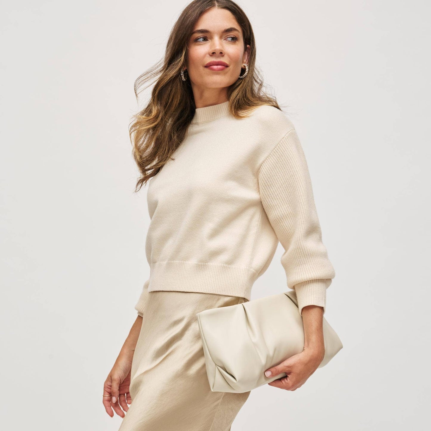 Woman wearing a cream sweater and pants holding a matching clutch against a white background