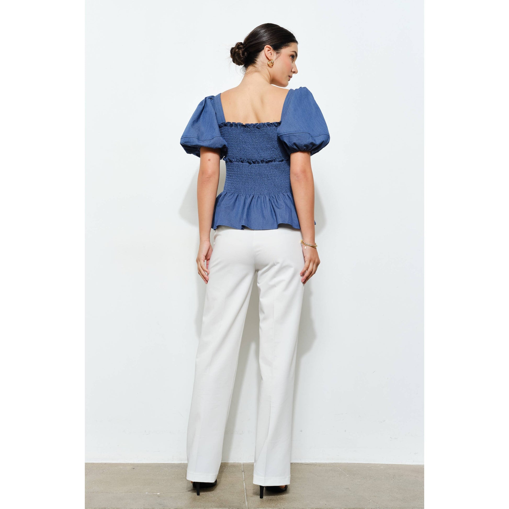 Woman wearing a blue top with puffed sleeves and white pants against a light gray background
