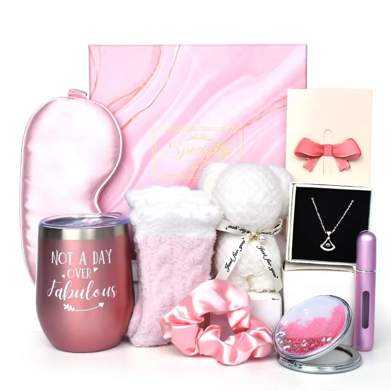 A luxurious pink-themed gift set featuring a silky sleep mask, stainless steel tumbler with 'Not a Day Over Fabulous' text, fuzzy pink-and-white socks, a plush teddy bear, a satin scrunchie, a dainty triangle pendant necklace in a gift box, a perfume atom