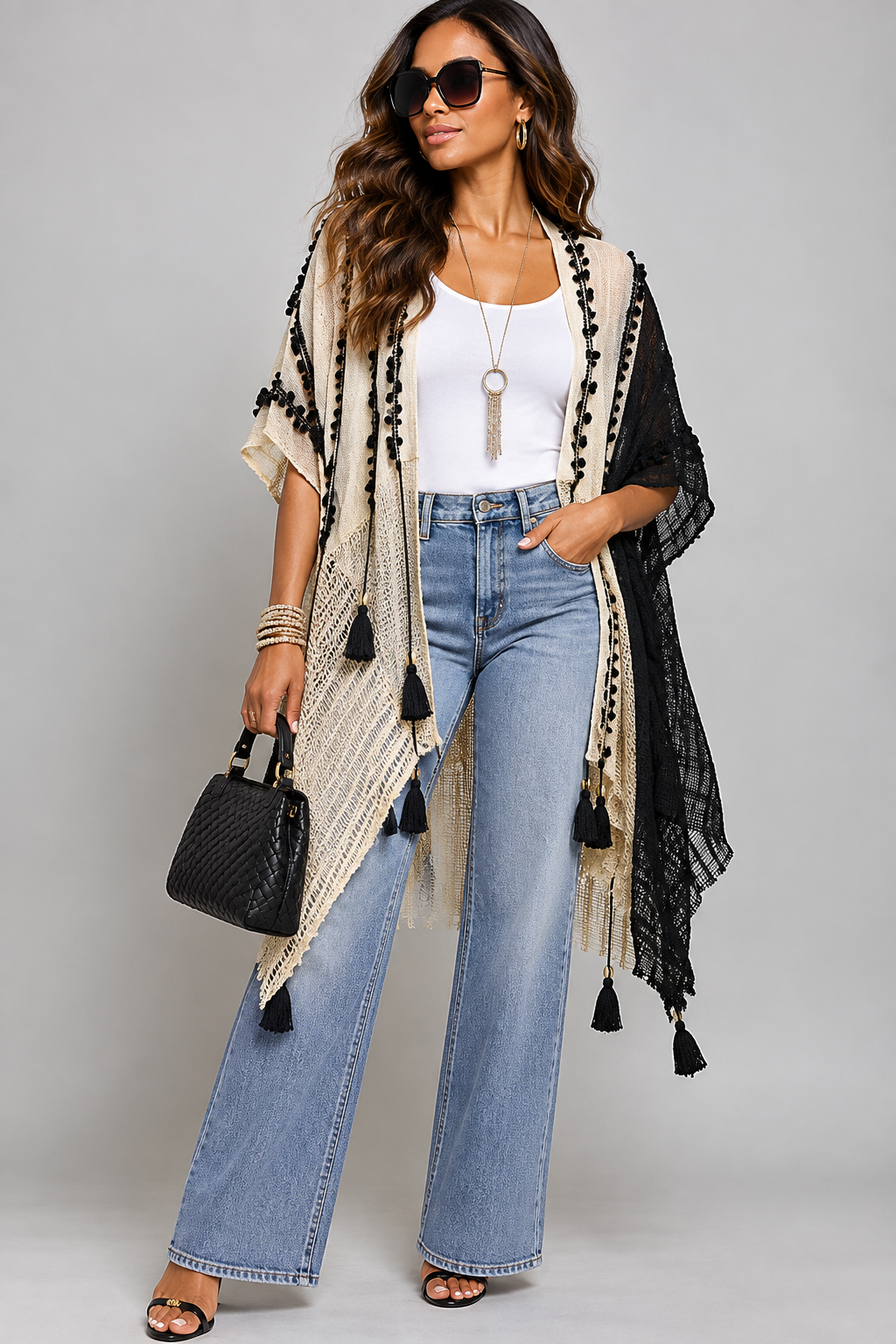 A woman models a black and cream knit open-front cardigan with tassel trim, worn over a white tank top and light-wash wide-leg jeans, against a solid gray studio background.