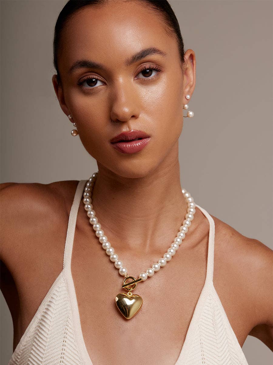 Woman wearing a pearl necklace with a gold heart pendant against a neutral background