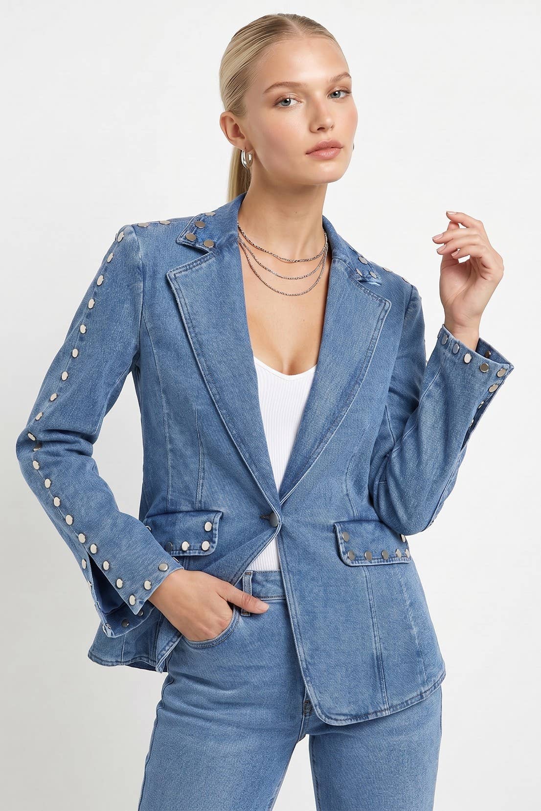 A woman modeling a blue denim blazer with silver button accents on the sleeves and lapels, paired with matching blue jeans and a white top, against a light gray studio background.