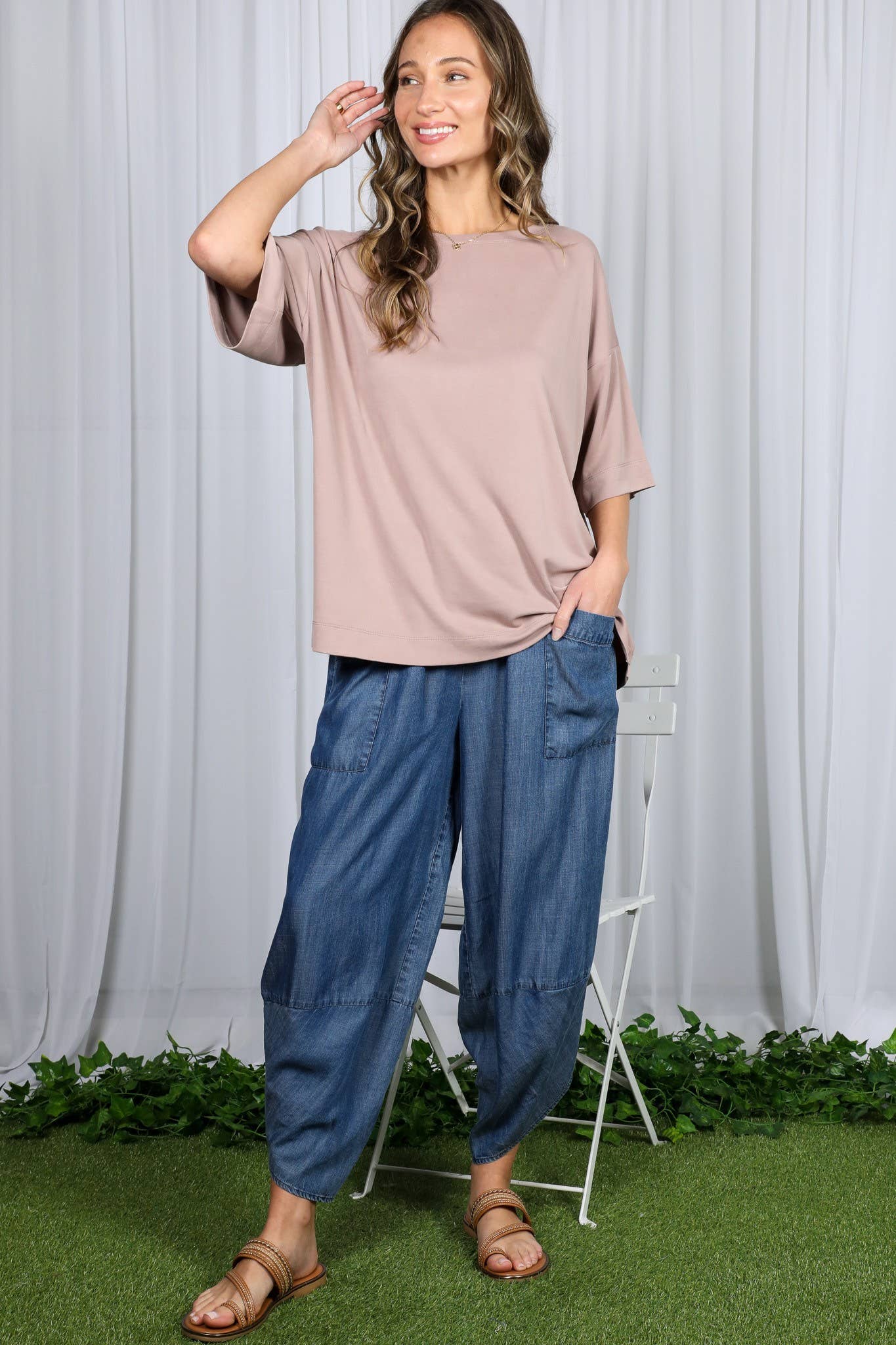 Woman wearing a beige top and blue pants standing against a white curtain background.